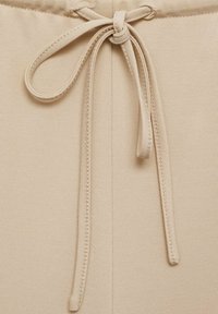 Beige fabric with a smooth texture featuring a tied drawstring detail at the waist, and contrasting stitched edges along the seams.