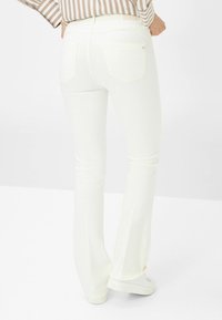 Weiße Schlagjeans mit glatter Textur, ausgestattet mit Gesäßtaschen und einer schmalen Passform bis zum Knie. Getragen mit weißen Sneakern.