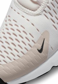 Ljusbeige atletisk sko med texturerat meshlager, med rosa accenter och en svart Nike-logotyp på en slät överdrag. Rund tå och flexibel sula.