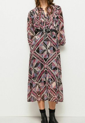 Frau trägt ein langes, gemustertes Kleid mit pinken, schwarzen und lila geometrischen Mustern, das mit einem schwarzen Gürtel tailliert ist, sowie schwarze Stiefeletten.