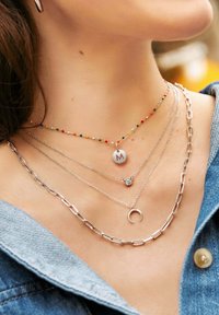 Le collane a strati presentano perline colorate, un ciondolo circolare con una "M", una catena d'argento, un dettaglio in cristallo e un'accentuata forma a mezzaluna.