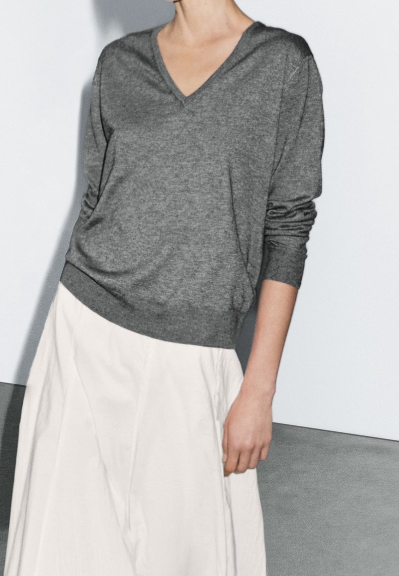 Personne portant un pull gris à col en V et manches longues, rentré dans une jupe taille haute de couleur écrue sur un fond uni.