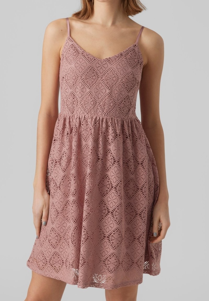 Femme portant une robe sans manches en dentelle rose poudré avec des motifs en losange et circulaires, corsage ajusté et jupe évasée.