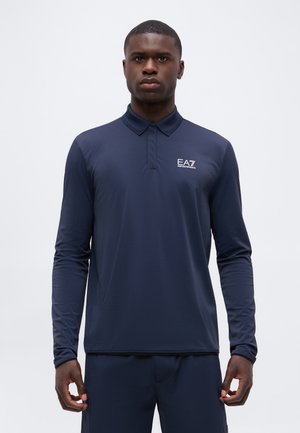 Mężczyzna ubrany w granatową koszulkę polo z długim rękawem z logo EA7 Emporio Armani na lewej piersi, stojący na jasnym, jednolitym tle.