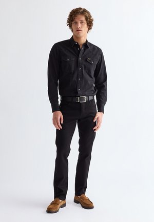 Νέος άνδρας με σγουρά μαλλιά στέκεται φορώντας μαύρο πουκάμισο με κουμπιά, μαύρο παντελόνι με ζώνη και καφέ μοκασίνια από σουέτ σε λευκό φόντο.