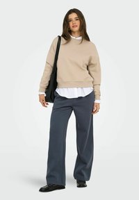 Beige sweatshirt med rund hals, lagd over en hvid skjorte med krave, parret med løse, mørkegrå bukser og sorte sko.