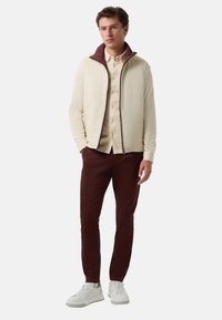 Herencrème jasje met bordeaux kraag, beige overhemd met knopen, bordeaux broek en witte sneakers. Gladde textuur, ritsluiting en contrasterende accenten.