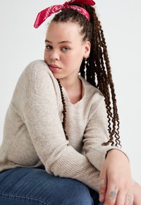 Beige ribbed sweater with a V-neck and long sleeves, paired with blue denim jeans. Red bandana accent in hair, featuring multiple ear piercings.