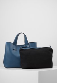 Modrá kožená taška přes rameno s texturovaným povrchem, vybavená dvěma rukojeťmi a kontrastním černým zipovým pouzdrem. Minimalistický design s čistými liniemi.