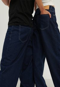 Zwei Personen stehen Rücken an Rücken und tragen dunkelblaue Jeans mit weitem Bein, eine mit der Hand in der Gesäßtasche, vor einem schlichten hellen Hintergrund.