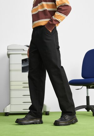 Pantalones negros en un diseño de pierna recta, usados con zapatos negros. El entorno incluye una alfombra verde y una impresora de oficina al fondo.