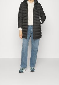 Chaqueta acolchada negra con capucha, con textura de acolchado horizontal, llevada sobre una camisa de color claro y jeans azules, combinada con zapatillas multicolores.