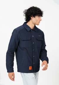 Donkerblauwe corduroy jas met een knoopsluiting aan de voorkant, twee borstzakken en een zak op de rechtermouw. Heeft een oranje merketiket aan de onderkant.