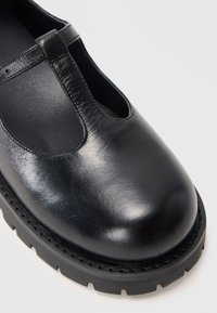 Black leather shoe with a rounded toe, T-bar strap, and a textured rubber sole featuring a zigzag pattern around the edge.