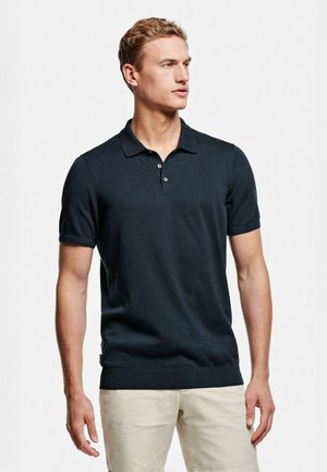 Jeune homme portant un polo noir à manches courtes et un short beige, debout et regardant vers la droite sur un fond blanc.
