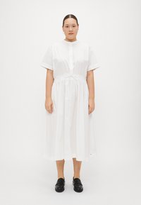 Weißes Baumwoll-Midikleid mit kurzen Ärmeln, durchgehender Knopfleiste und Bindegürtel in der Taille. Trägt man mit schwarzen Slippern vor einem neutralen Hintergrund.