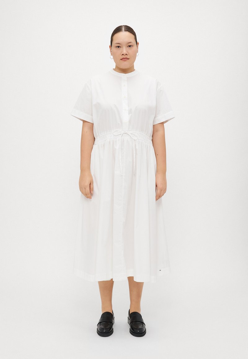 Weißes Baumwoll-Midikleid mit kurzen Ärmeln, durchgehender Knopfleiste und Bindegürtel in der Taille. Trägt man mit schwarzen Slippern vor einem neutralen Hintergrund.