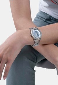 Srebrny zegarek z okrągłą tarczą i metalową bransoletą. Niebieska tarcza ma błyszczącą teksturę i przezroczyste kamienie wokół pierścienia. Widoczny okrągły detal korony.