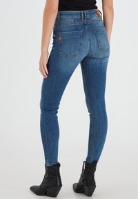 Tievās džinsās tumšā zilos denimā ar gludu tekstūru, augsto jostasvietu, divām aizmugures kabatām un ādas jostas akcents. Kombinētas ar melniem potīšu zābakiem.