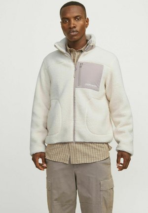 Fleece jakna v off-white barvi z zadrgo na prsnem žepu, sivi poudarki in dvema sprednjima žepoma. Teksturirana, mehka površina z visokim ovratnikom.