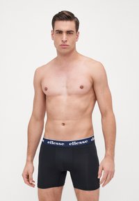 Black boxer shorts made of soft fabric, featuring a navy waistband with white "ellesse" branding, fitted design, and mid-thigh length.