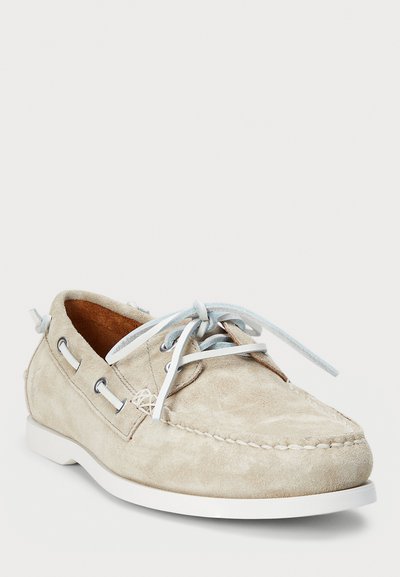 Beige suede boat shoe with white rubber sole, blue laces, and silver eyelets. Features moccasin stitching and a soft, textured surface.
