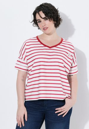 Kvinde med kort krøllet hår iført en hvid t-shirt med røde vandrette striber og blå jeans, stående med den ene hånd i lommen.