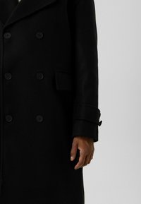 Zwarte dubbele rij knopen jas met zes knopen, klepzak en banddetail op de mouw, gedragen door een persoon met zichtbare rechterhand.