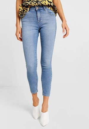 Kobieta ubrana w jasnoniebieskie obcisłe spodnie, żółto-czarną kwiecistą bluzkę oraz białe szpilki z noskiem w szpic, stojąca na białym tle.