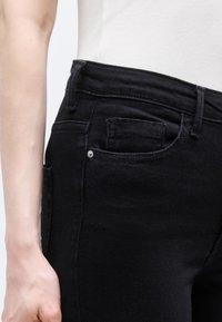 Κοντινό πλάνο ενός ατόμου που φοράει μαύρο τζιν και λευκό ριμπ μπλουζάκι, εμφανίζοντας τον γοφό, την μπροστινή τσέπη και το μέρος του βραχίονα.