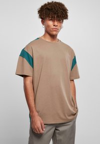 Beige T-Shirt in lässiger Passform mit türkisfarbenen Akzenten auf den Schultern, kurzen Ärmeln und einem runden Ausschnitt. Weicher, glatter Stoff mit minimaler Textur.