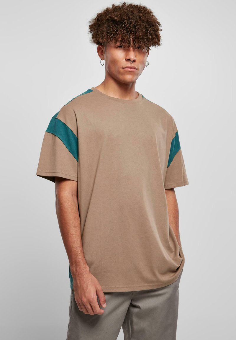 Beige T-Shirt in lässiger Passform mit türkisfarbenen Akzenten auf den Schultern, kurzen Ärmeln und einem runden Ausschnitt. Weicher, glatter Stoff mit minimaler Textur.