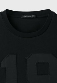 T-shirt noir à col rond avec un motif en relief du numéro 19 sur le devant et une étiquette de la marque Stradivarius à l'intérieur du col.