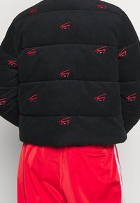 Czarna kurtka puchowa z sztruksu z czerwonymi akcentami logo, pikowanym wzorem i pudełkowym fasonem. Połączona z jasnoczerwonymi spodniami.