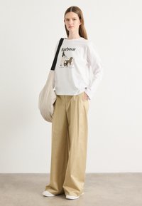 Chemise blanche à longues manches avec imprimé et illustration "Barbour", associée à un pantalon large beige. Le modèle porte un sac beige et des chaussures blanches.