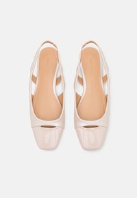 Une paire de ballerines beige à bride arrière avec un bout carré en vernis et un détail découpé, vue de dessus sur un fond blanc.