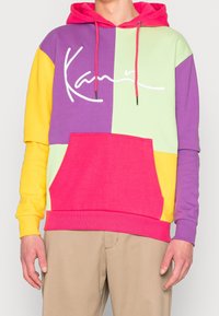 Sudadera con capucha colorida en secciones moradas, verdes, amarillas y rosas, con un bolsillo frontal y un logo blanco. Tejido texturizado, diseño casual.