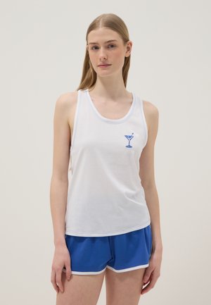 Young woman wearing white sleeveless tank top with blue cocktail glass design and blue athletic shorts against plain background.