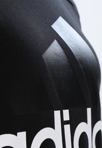 Zwarte stof met een gladde textuur met een groot wit Adidas-logo. Boven het logo zijn drie geometrische vormen in het grijs zichtbaar.