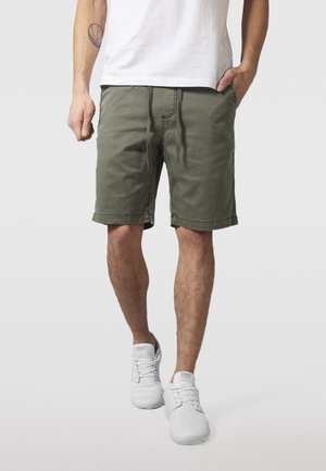 Homme portant un short vert olive avec cordon de serrage, un t-shirt blanc et des baskets blanches, se tenant avec une main dans sa poche devant un fond uni.