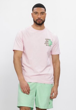 Rövid hajú és szakállas férfi világos rózsaszín pólóban, amelyen szöveg és zöld grafika látható, világos zöld rövidnadrággal, fehér háttér előtt áll.