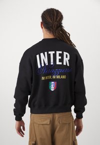 Crna majica sa kapuljačom s velikim natpisom "INTER", plavim detaljima i zlatnim slovima. Stranica s leđa uključuje grafički emblem s šarenim štitom.