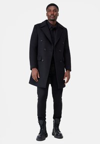 Svart dubbelknäppt kappa i en ullblandning med spetsad krage, sidfickor och texturerad yta, kombinerad med åtsittande svarta byxor och stövlar.