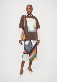 Brązowa, oversize koszulka z grafiką kota, nałożona na jasnoróżową koszulę. Kolorowa, abstrakcyjnie wzorzysta spódnica midi i białe szpilki.