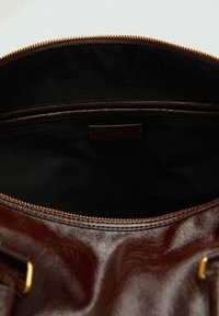 Sac en cuir marron foncé avec fermeture éclair dorée, ouvert pour révéler un intérieur en tissu noir et une poche intérieure.