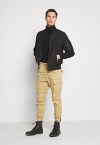 Svart bomberjacka med texturerade ärmar, åtsittande svart polotröja, beige cargobyxor med sidofickor och svarta snörade kängor.
