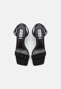 Sorte ruskinds hæle sandaler med firkantet tå, ankelrem og en glat, blank indersål. Har en bred frontrem for støtte.