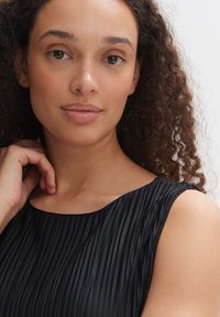 Black pleated dress with a round neckline, soft texture, and shoulder straps. Model has curly hair and lightly tinted natural makeup.