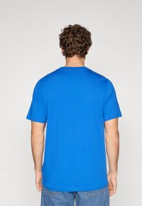 Persona ar cirtainiem matiem, valkājot vienkāršu zilu īsroku krekliņu un zilas džinsa bikses, redzama no muguras uz gaiša fona.