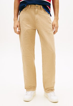 Uomo che indossa pantaloni beige con trama, sneakers bianche e una maglietta a righe rossa, blu navy e bianca, mostrato dalla vita ai piedi.